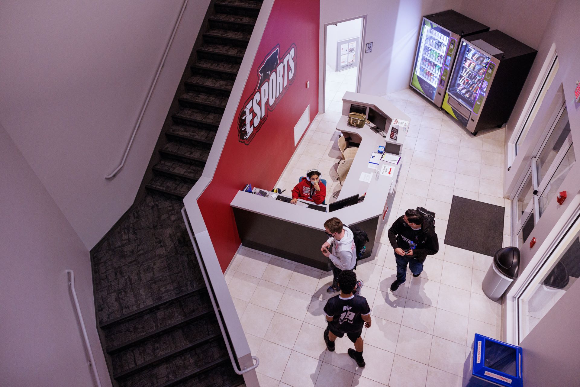 Aerial view of the entrance area of the Esports center featuring a reception desk with a staff member, vending machines, and students standing and interacting